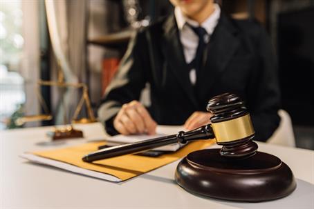 Close-up of a judge's gavel on a desk with legal documents, scales of justice, and a lawyer in the background.